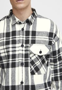 Camicia di flanella a quadri bianca e nera, con frontale a bottoni, colletto a punta e tasca sul petto con un solo bottone. Texture morbida.