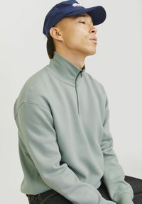 Light green zip-up sweatshirt with a high collar, made of smooth fabric. Paired with a navy cap featuring a logo. Black jeans visible.