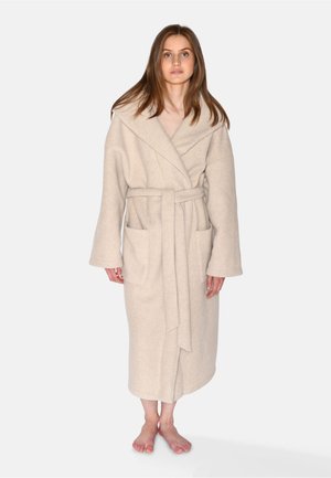 Femme aux longs cheveux bruns, debout pieds nus, portant un manteau en laine beige ceinturé avec grand col et poches avant.