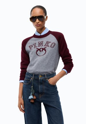 Donna con occhiali da sole tartarugati, maglione Pinko bordeaux e grigio, camicia blu, jeans a vita alta e portachiavi a forma di fiore.