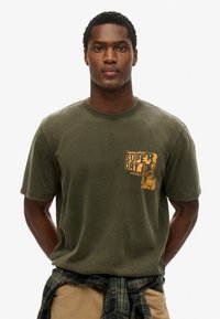 Oliwkowa bawełniana koszulka z okrągłym dekoltem, z żółtym nadrukiem na lewej piersi. Miękka tekstura, luźny krój.