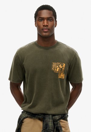 Camiseta de algodón verde oliva con cuello redondo, que presenta un estampado gráfico amarillo en el lado izquierdo del pecho. Textura suave, ajuste casual.