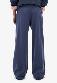 Persona de pie de espaldas que lleva unos pantalones azules anchos y holgados, una sudadera azul marino y los puños de una camisa rosa visibles.