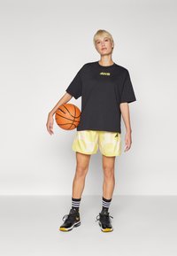 Must have kašmiri t-särk, millel on kollane logo, kollase mustriga lühikesed püksid, triibulised mustad ja valged sokid ning mustad sportlikud kingsad, pidades käes korvpalli.