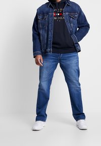 Denim jacket over a navy hoodie with a logo, paired with light blue jeans and white sneakers, showcasing a casual layered outfit.