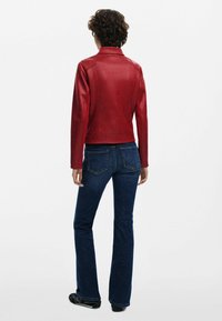 Rote Kunstlederjacke mit taillierter Passform und Stehkragen, kombiniert mit dunkelblauen Schlagjeans und schwarzem Schuhwerk.
