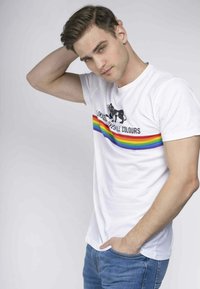 Hvid bomuld t-shirt med et sort grafisk print og en vandret regnbuestribe over brystet. Bæres med blå denim jeans.