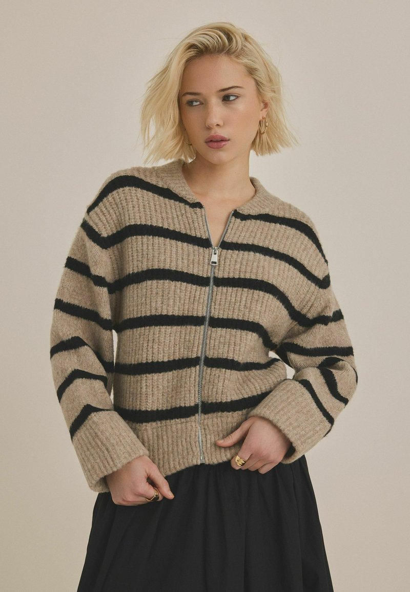 Beige randig cardigan med svarta horisontella ränder, ribbad textur och dragkedja framtill. Har breda ärmslut och en rundad krage.