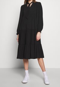 Robe noire à volants avec des longues manches et un décolleté en trou de serrure, associée à des baskets blanches. Le tissu semble texturé et doux.