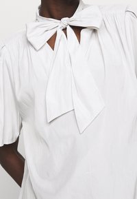 Bílá halenka vyrobená z lehkého materiálu, s vázanou mašlí na krku, plisovanými ramenními částmi a krátkými balónovými rukávy. Hladký povrch.