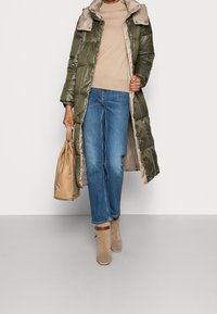 Un abrigo acolchado largo en verde oliva con detalles en beige, combinado con una blusa beige, jeans azules y botines beige. Incluye un bolso marrón.