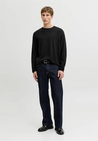 Jeune homme debout portant une chemise noire à manches longues, un jean bleu foncé, une ceinture noire et des chaussures en cuir noires, sur un fond clair uni.