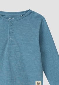 Blauw overhemd met lange mouwen van zachte stof, voorzien van een drieknoopsluiting; heeft een gestructureerd oppervlak en een klein geborduurd embleem aan de zoom.