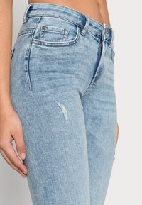 Jeans de denim azul claro com uma cintura alta, apresentando um remendo desgastado na coxa, cinco bolsos e desbotamento subtil por todo o lado.