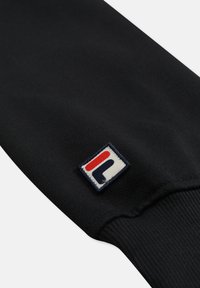 Zwarte stoffen mouw met geribde manchetten en een klein vierkant lapje met een rood- en marineblauw geometrisch logo.