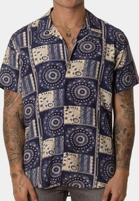 Camisa de botones de manga corta con un diseño de patchwork en azul marino y crema, con patrones circulares, tejido texturizado y un ajuste relajado.