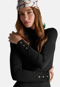 Čierny top s dlhými rukávmi, ktorý má zlaté gombíky na manžetách, hladkú textúru a priliehavý strih. Na hlave má vzorovaný šál.