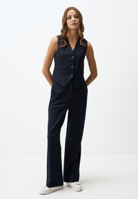 Marineblaue, taillierte Weste mit fünf Knöpfen, kombiniert mit passgenauen, weiten Hosen und weißen Sneakers. Das Material hat eine glatte Textur.