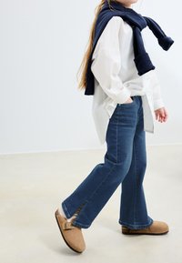 Biela košeľa, modré rozšírené denimové džínsy, hnedé semišové dreváky s prackami a tmavomodrý sveter prehodený cez plecia. Jednoduché pozadie.