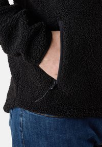 Svart fleece-pull med texturerad yta, utrustad med ett dragkedjefack på sidan och elastiska cuffs, i kombination med blå denimjeans.