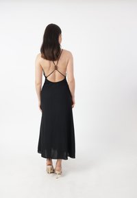 Schwarzes, rückenfreies Maxikleid mit dünnen Trägern und einem dekorativen Akzent am Rücken. Glatter Stoff, der bis zum Boden fließt, kombiniert mit beigen Absätzen.