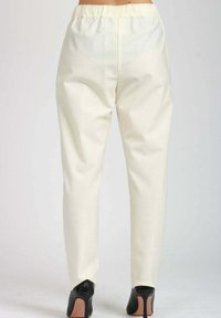 Donna che indossa pantaloni crema a vita alta, dal taglio morbido e con cintura elastica, abbinati a scarpe col tacco nere, vista da dietro.