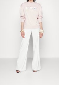 Světle růžová mikina s grafikou "HUGO 1983", v kombinaci s širokými bílými kalhotami. Doplněno o naskládané náramky a černé sandály na podpatku.