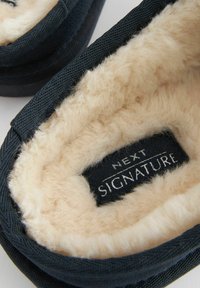 Crne platnene papuče s mekim bež umjetnim krznom iznutra i crnom oznakom sa natpisom "NEXT SIGNATURE" ušivenom unutra.