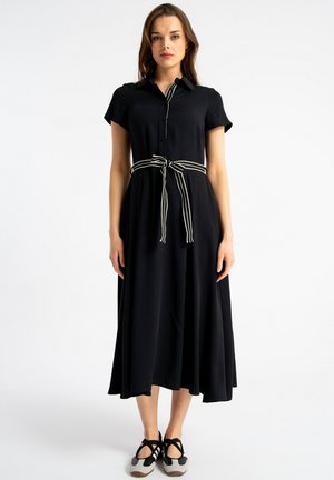 Kobieta w czarnej sukience midi z krótkim rękawem i białym paskiem w paski zawiązanym w talii, stoi w czarno-białych butach na białym tle.