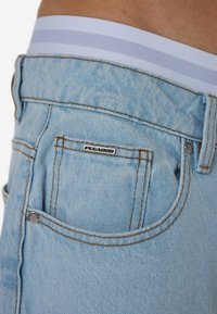 Ανοιχτό μπλε τζιν με πορτοκαλί ραφές, που διαθέτει πίσω τσέπη με μεταλλική λεπτομέρεια με την ένδειξη "PEGAOOR" και λευκή μέση με γκρι ρίγες.