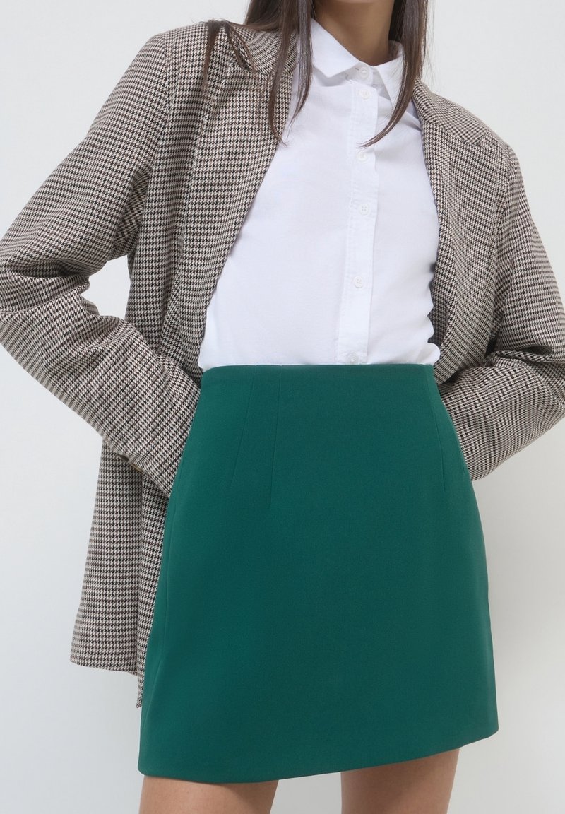 Femme portant une chemise blanche boutonnée, un blazer marron à carreaux et une jupe verte taille haute, les mains dans les poches du blazer.