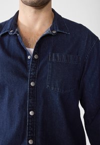 Mörk denimskjorta med knapplås framtill, featuring metallsnappar, en vänster bröstficka och en klassisk kragsdesign.