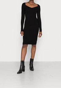 Robe noire moulante côtelée à manches longues et col carré, associée à des bottines en cuir noir à talon carré.