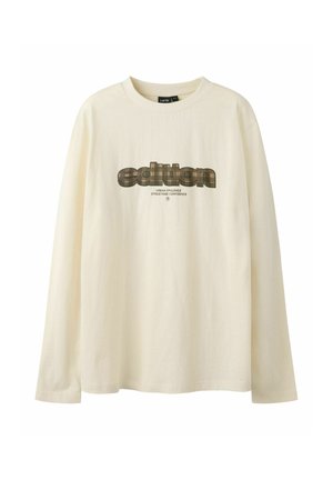Camiseta de manga larga color crema con cuello redondo y la palabra "edition" en fuente de cuadros sobre un texto más pequeño que dice "urban opulence streetwise confidence."