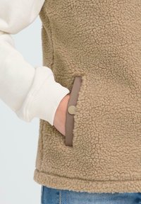 Main dans la poche d'un gilet en sherpa beige porté par-dessus une chemise à manches longues blanche avec des poignets côtelés et un jean bleu.