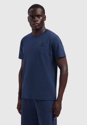 Ein Mann steht und trägt ein marineblaues kurzärmeliges Hemd und passende Shorts mit dezenten aufgedruckten Logos auf Brust und Oberschenkel.