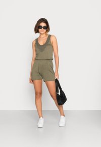 Camisola verde-azeitona sem mangas com decote decorativo, calções a condizer e ténis brancos. Bolsa preta de ombro adiciona contraste. Fundo simples.