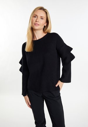 Sort strikket sweater med flagrende volanger på ærmerne. Har en rund halsudskæring og tekstureret stof, parret med sorte bukser.