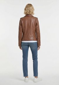 Bruine leren jas met aangesloten pasvorm, ritssluitingen aan de manchetten en een spitse kraag. Gecombineerd met een blauwe spijkerbroek en witte sneakers, gezien van achteren.