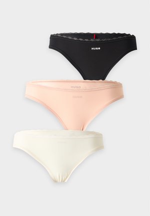 Femme debout pieds nus sur fond blanc, portant un soutien-gorge beige clair et une lingerie assortie, faisant face à l’avant avec les mains jointes devant.