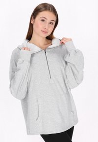Šedá zipová mikina s klokaní kapsou, měkká tkanina, oversized střih a kontrastní bílá podšívka kapuce. Obsahuje manžetové rukávy.