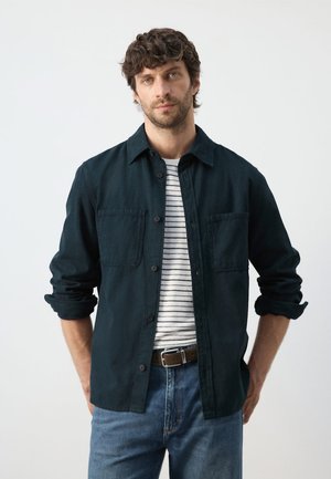 Homme aux cheveux bouclés portant une chemise noire boutonnée sur un t-shirt blanc à rayures noires, jean bleu et ceinture marron, debout les mains derrière le dos.