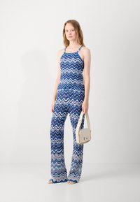 Une combinaison à motifs chevrons bleus et blancs avec des bretelles spaghetti, des pantalons évasés et un corsage ajusté. Associée à un petit sac à main crème.