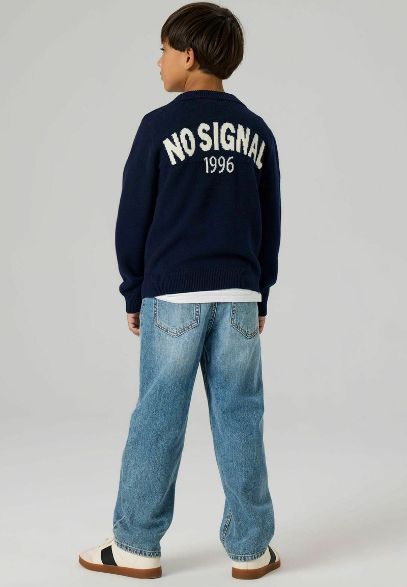 Marineblauer Pullover mit der Aufschrift „NO SIGNAL 1996“ auf der Rückseite, kombiniert mit hellblauen Jeans und weißen Schuhen mit schwarzem Akzent. Weiche Strickstruktur.