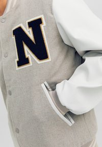 Grijs en witte varsityjas met een geborduurd zwart "N" logo, getextureerd wollen lichaam, leren mouwen, drukknopen en ribgebreide manchetten.