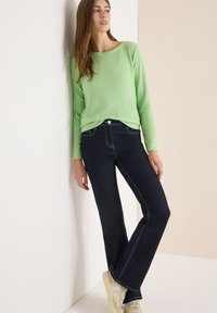 Grüner, gerippter Pullover mit langen Ärmeln, dunkelblaue ausgestellte Jeans mit kontrastierenden Nähten und beige Sneaker. Stehend vor hellem Hintergrund.