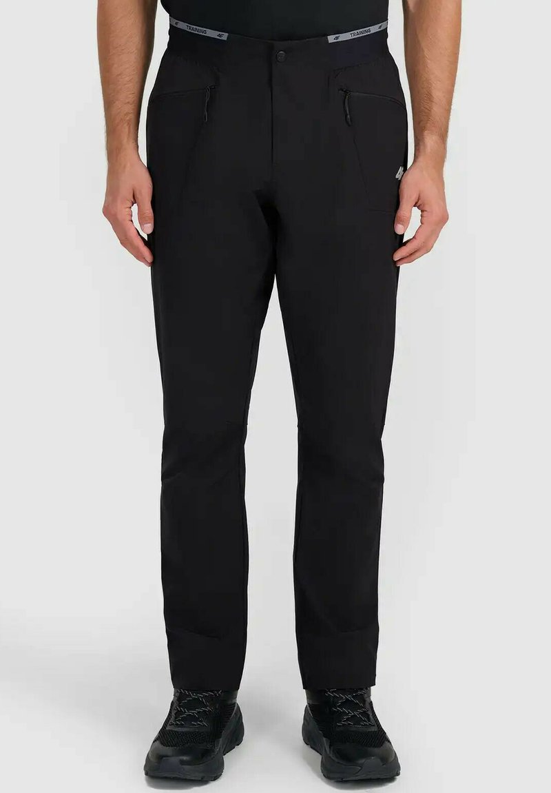 Homme portant un pantalon noir slim avec poches avant zippées et chaussures de sport noires, debout devant un fond uni.