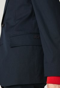 Marinblå skräddarsydd kavaj med en slät yta, som har en knapp, sidofickor och en broderad logotyp ovanför fickan.