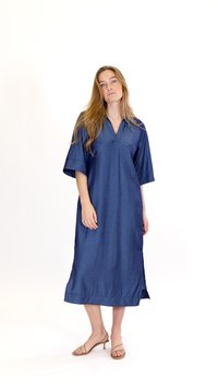 Naine, kellel on pikad blondid juuksed, kannab lahtist sinist midi kleiti ja beeže rihmlukkudega kontsakingi, seisab valgel taustal.