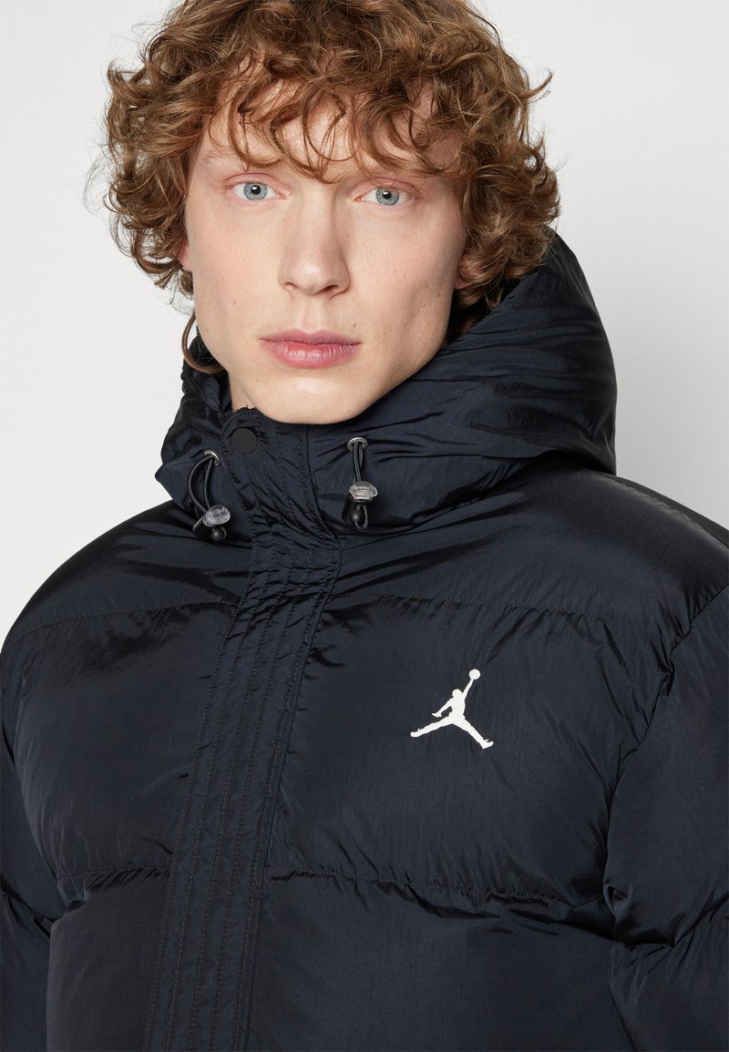 Młoda osoba z kręconymi włosami, nosząca czarną kurtkę puchową z kapturem i białym logo Jumpman na piersi.